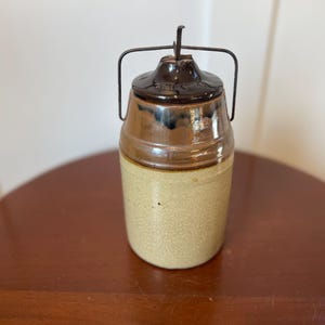 May include: A brown and tan ceramic jar with a metal lid and handle. The jar has a speckled glaze and a dark brown rim.