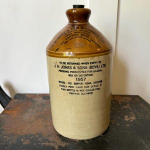 May include: A vintage stoneware jug with a brown top and a tan body. The jug has a dark brown stopper and printed text. The text includes the company name, address, and the year 1957. The jug is on a dark surface.