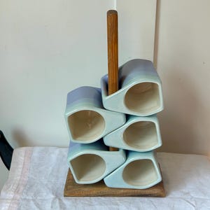 May include: A wooden mug tree stand holding five ceramic mugs. The mugs feature a light blue and white gradient glaze with a smooth finish. The stand has a square wooden base and a central vertical wooden post that supports the stacked mugs.
