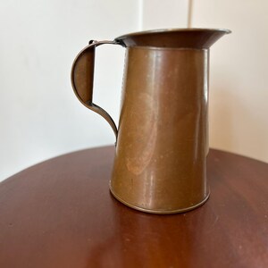 May include: A small, antique copper pitcher with a curved handle and a flared spout. The pitcher is a warm, reddish-brown color and sits on a dark wooden surface. The pitcher is likely used for serving liquids.