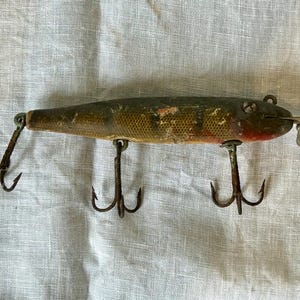 May include: An antique fishing lure with a weathered appearance. The lure is primarily olive green and tan, with red accents near the front. It features three treble hooks and a small propeller at the front, indicating its use for attracting fish.