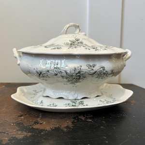 May include: A white porcelain soup tureen with a lid, handles, and a matching serving plate. The tureen and plate are decorated with a green floral pattern. The tureen is sitting on a dark surface.
