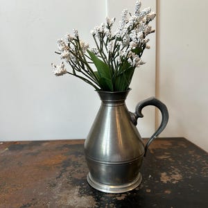 May include: A metallic pewter-colored pitcher with a rounded base, a tapered neck, and a curved handle. The pitcher contains a bouquet of artificial white flowers with small, textured buds and green leaves, creating a contrast in color and form.