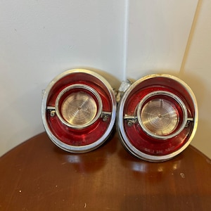 May include: Pair of vintage car tail lights. Each light features a red lens with a chrome bezel. The center has a silver, spiral-patterned reflector. The lights are mounted on a metal backing.