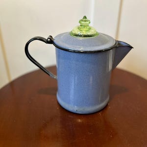 May include: A vintage blue enamelware coffee pot with a black handle and spout. The lid is gray with a green glass knob. The pot is sitting on a round, brown wooden table.