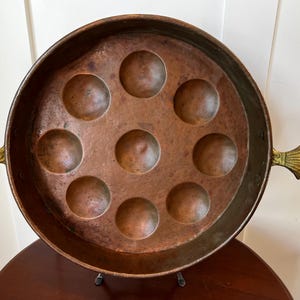 May include: A copper pan with eight round indentations and two brass handles shaped like seashells. The pan is on a wooden stand.