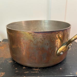 May include: A round copper cooking pot with a metallic interior and a single brass handle attached with two rivets. The exterior surface shows a weathered, tarnished patina with visible dark spots and variations in the warm copper-toned finish.