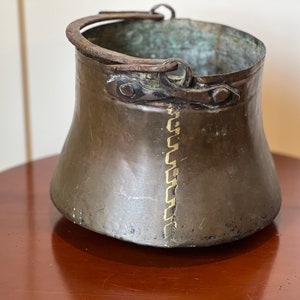 May include: A vintage copper bucket with a metal handle. The bucket has a decorative gold design around the top and a seam down the side.