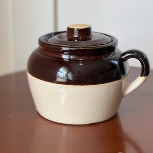 May include: A small brown and white ceramic crock pot with a lid and a handle. The crock pot is glazed and has a rustic look.