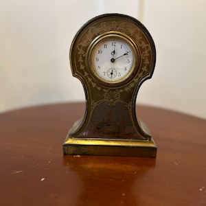 May include: An antique brass-toned mantel clock with a white clock face and black numerals. The clock has an ornate design with floral and scrollwork details. The clock is set on a wooden table.