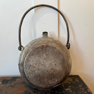 May include: A weathered, round metal pot with a curved handle. The pot has a dark, aged appearance with visible texture and a small spout. The handle is attached with metal rings.