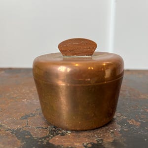 May include: A small, copper-colored container with a matching lid and a wooden handle. The container has a rounded shape and sits on a dark, textured surface. The lid is a small, rectangular piece of wood.