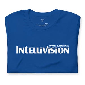 May include: A royal blue t-shirt with a white Intellivision logo. The logo reads "Mattel Electronics Intellivision".