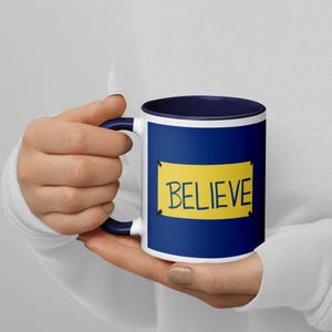 Op de afbeelding: Een witte keramische mok met een marineblauwe buiten- en binnenkant. De mok heeft een gele rechthoek met het woord "BELIEVE" in zwarte, handgeschreven letters. De mok wordt door een persoon vastgehouden.