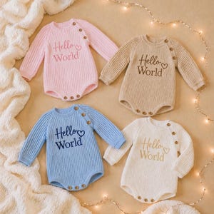 May include: Four knitted baby rompers in pastel colors: pink, beige, blue, and white. Each romper has long sleeves, wooden buttons, and the embroidered text "Hello World" with a heart symbol. The rompers are arranged on a soft, textured surface.