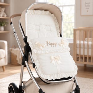 May include: A cream-colored stroller footmuff featuring a ruffled border, three decorative bows, and the name Poppy embroidered in brown script. The padded fabric insert is designed to fit into a stroller seat with a silver metal frame.