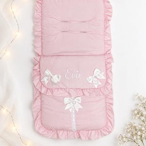 May include: A rectangular, light pink padded stroller liner featuring a ruffled border, three white satin bows, and a vertical lace trim. The name Evie is embroidered in white script on the center flap, which also includes two horizontal stitched slots.