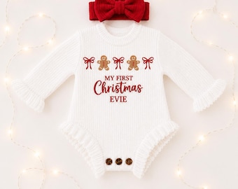 Könnte beinhalten: Ein weißer, langärmeliger Strickstrampler mit einem roten Schleifen-Stirnband. Der Strampler ist mit Lebkuchenmännern und roten Schleifen verziert und trägt den roten Schriftzug "MY FIRST Christmas EVIE" sowie drei braune Knöpfe am Saum.