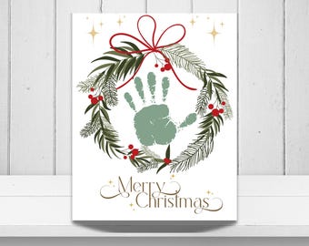 May include: A white rectangular print featuring a central sage green handprint surrounded by a circular wreath of dark green pine needles and red berries. A red bow sits at the top of the wreath. The text Merry Christmas is printed in gold.