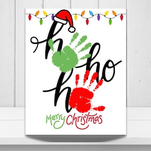May include: A white rectangular print featuring the black cursive text Ho Ho Ho with a red and white Santa hat on the first letter. A green handprint replaces the first o and a red handprint replaces the second o. Multi-colored lights and Merry Christmas text.