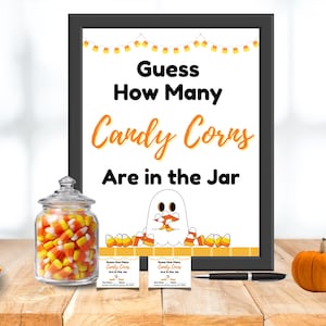 May include: A Halloween-themed display featuring a framed sign that reads "Guess How Many Candy Corns Are in the Jar." A glass jar filled with candy corn, two small cards, a pen, and two pumpkins are on a wooden surface.