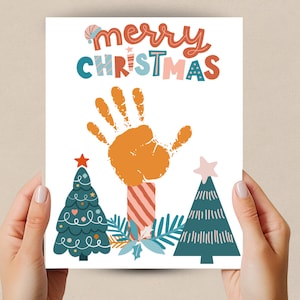 Könnte beinhalten: Eine weiße Karte mit dem Text "merry christmas" in bunten Buchstaben. Ein orangefarbener Handabdruck befindet sich in der Mitte der Karte, mit zwei grünen Weihnachtsbäumen auf jeder Seite des Handabdrucks.