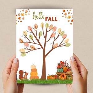 Puede incluir: Una impresión con temática otoñal con un árbol con hojas coloridas, calabazas y una ardilla. Las palabras "hello FALL" están escritas en la parte superior. La impresión está sostenida por dos manos.