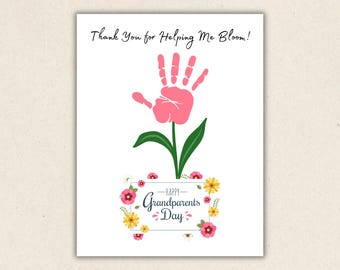 Puede incluir: Una tarjeta de felicitación blanca con una huella de mano rosa en forma de flor, con tallo y hojas verdes. El texto dice Thank You for Helping Me Bloom! en la parte superior y Happy Grandparents Day dentro de un marco rectangular con flores.
