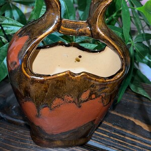 May include: A brown and orange ceramic planter with a unique, abstract design. The planter has a wide opening and two handles on the top.
