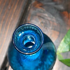 Puede incluir: Una botella de vidrio azul con una boca ancha y una base redondeada. La botella tiene una superficie texturizada y una abertura clara en la parte superior.