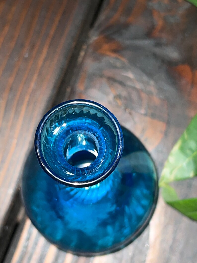 Puede incluir: Una botella de vidrio azul con una boca ancha y una base redondeada. La botella tiene una superficie texturizada y una abertura clara en la parte superior.