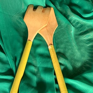 May include: A pair of wooden salad servers with yellow handles. The servers are crossed and lying on a green fabric background.