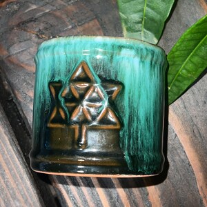 May include: A small, dark brown ceramic mug with a green glaze. The mug has a geometric design on the front, resembling a star or a flower.