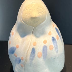 May include: A ceramic figurine of a person in a hooded garment. The figure has closed eyes and a gentle smile. The off-white figure is decorated with blue and orange abstract shapes.