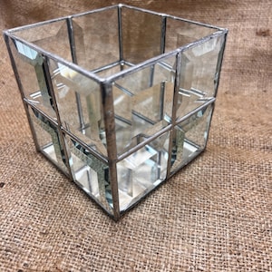 May include: A clear glass cube-shaped decorative object with a silver metal frame. The glass panels are faceted, creating a multi-dimensional effect. The object is sitting on a brown burlap surface.