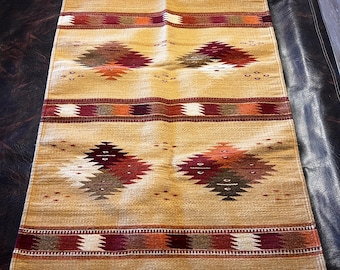 Puede incluir: Una alfombra rectangular tejida con un fondo beige y patrones geométricos de diamantes en tonos de rojo, naranja y marrón. La alfombra tiene flecos blancos en los extremos y rayas horizontales.