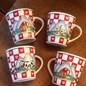 May include: Set of four white ceramic mugs with red and white checkered pattern and mountain scenes. Each mug features a different scene: a cow, a person in a red and yellow outfit, a person in a blue and yellow outfit, and a flower.