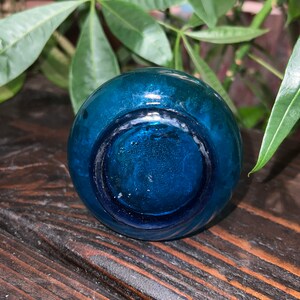 Puede incluir: Una botella de vidrio azul verdoso con fondo redondo. La botella est&aacute; sobre una superficie de madera oscura.