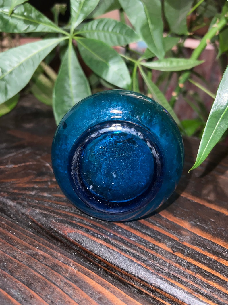 Puede incluir: Una botella de vidrio azul verdoso con fondo redondo. La botella est&aacute; sobre una superficie de madera oscura.