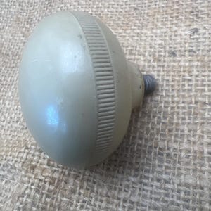 Può includere: Un pomello ovale verde chiaro con una fascia a coste al centro. Una vite metallica è attaccata alla base. Si tratta probabilmente di una maniglia per armadietto o cassetto.