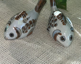 Puede incluir: Dos figuritas de cerámica de peces con diseños pintados en marrón y blanco. Los peces están mirando en direcciones diferentes y tienen ojos negros.