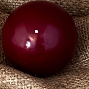 May include: A glossy, deep red sphere with a small white dot, resting on a textured, tan fabric. The smooth surface of the sphere reflects light, creating highlights. The object appears to be a decorative element or a component of a larger item.