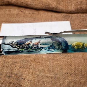 May include: A clear pen with a detailed landscape design inside. The design features seals on a rocky shore with a blue ocean and a yellow object. The pen has a silver clip.