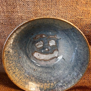 May include: A blue ceramic bowl with a white glaze and a brown rim. The bowl has a unique design with a smiley face in the center.