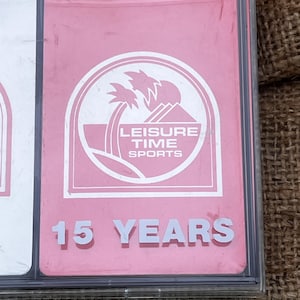 May include: A pink card with the words "LEISURE TIME SPORTS" inside a white circle with palm trees. The text "15 YEARS" is below the logo. The card is in a clear plastic case.