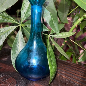 Puede incluir: Un peque&ntilde;o jarr&oacute;n de vidrio azul verdoso con cuerpo redondeado y cuello estrecho. El jarr&oacute;n est&aacute; sobre una superficie de madera oscura.