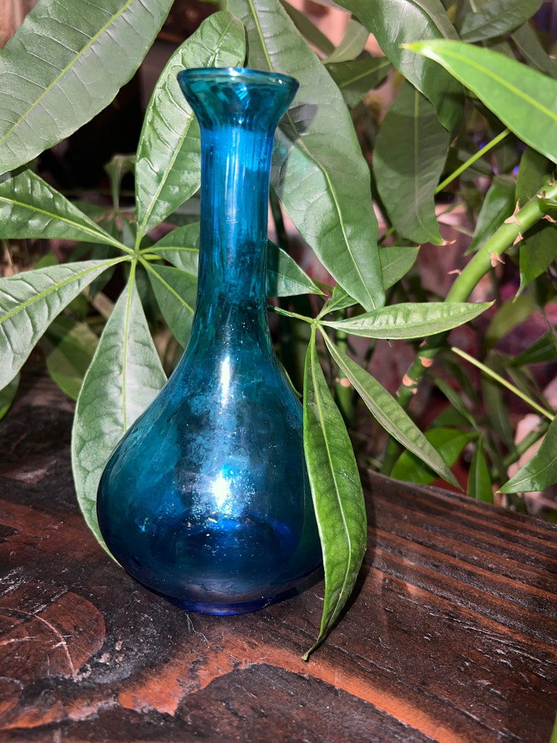 Puede incluir: Un peque&ntilde;o jarr&oacute;n de vidrio azul verdoso con cuerpo redondeado y cuello estrecho. El jarr&oacute;n est&aacute; sobre una superficie de madera oscura.