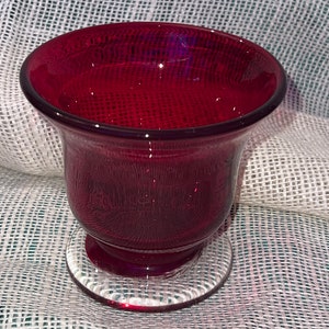 May include: A deep red glass vase with a flared top and a clear glass base. The vase is sitting on a textured fabric surface.