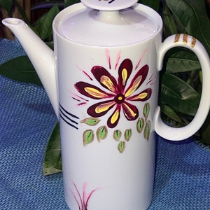May include: White ceramic teapot with a floral design. The teapot has a lid with a green leaf design and a gold trim around the handle. The floral design features a large red and yellow flower with green leaves and pink accents.