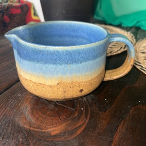 May include: A blue and brown ceramic pitcher with a handle. The pitcher has a glazed finish and a speckled texture.